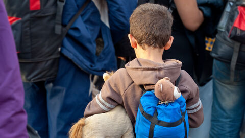 Im Bildvordergrund zu sehen ist ein kleiner Junge mit Kuscheltier und Rucksack, im Bildhintergrund Erwachsene.