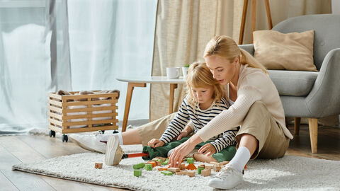 Kind sitzt mit ihrer Mutter auf einem Teppich und spielt mit Holzklötzen.