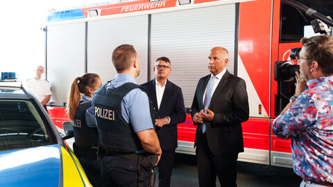 Ministerpräsident Rhein und Innenminister Poseck stehen vor einem Feuerwehrfahrzeug und unterhalten sich mit zwei Polizisten.