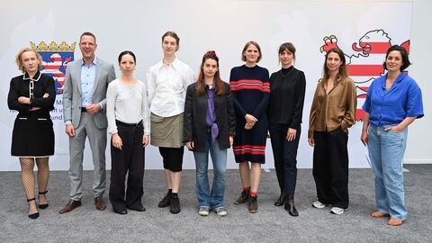 Staatssekretär Degen mit acht Frauen stehen in einer Reihe. Auf der Wand hinter ihnen ist das Hessen Wappen aufgedruckt.
