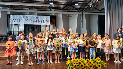 Viele Schülerinnen und Schüler halten Sonnenblumen in den Händen. Auf einem Banner steht "Herzlich Willkommen"