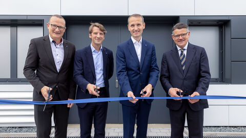 Vier Männer stehen in Anzügen gekleidet nebeneinander. Vor ihnen ist ein blaues Band gespannt, das sie mit einer Schere durchschneiden wollen. 