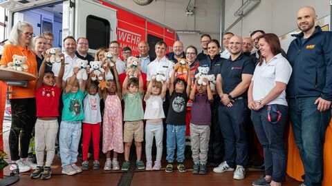 Innenminister Poseck steht gemeinsam mit mehreren Erwachsenen in einer Garage der Feuerwehr. Vor ihnen stehen auch Kinder, die einen Teddy hochhalten.